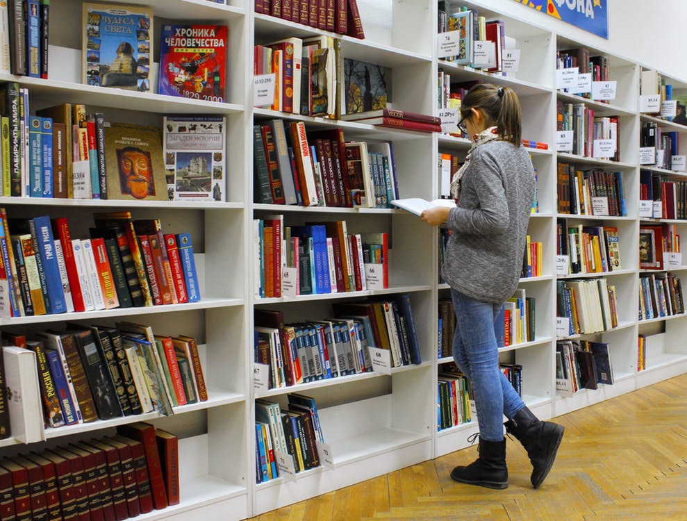 noor naine seisab suure raamaturiiuli ees avatud raamat käes