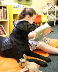 Tüdruk istub koos musta retriiveriga raamatukogu põrandal suurel padjal ja loeb raamatut.